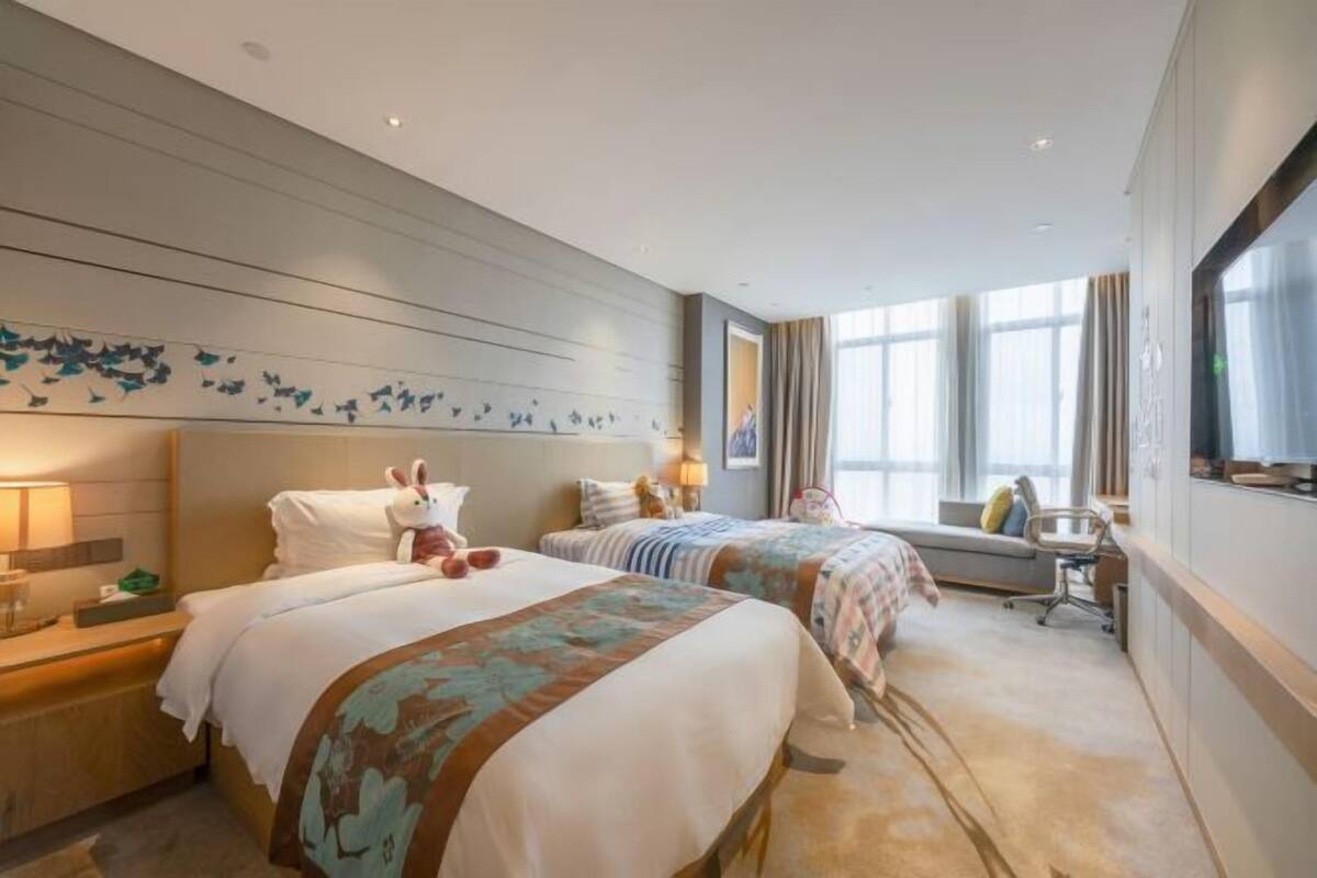 Quarto de hotel com duas camas, uma janela grande, uma obra de arte na parede e uma escrivaninha com cadeira.