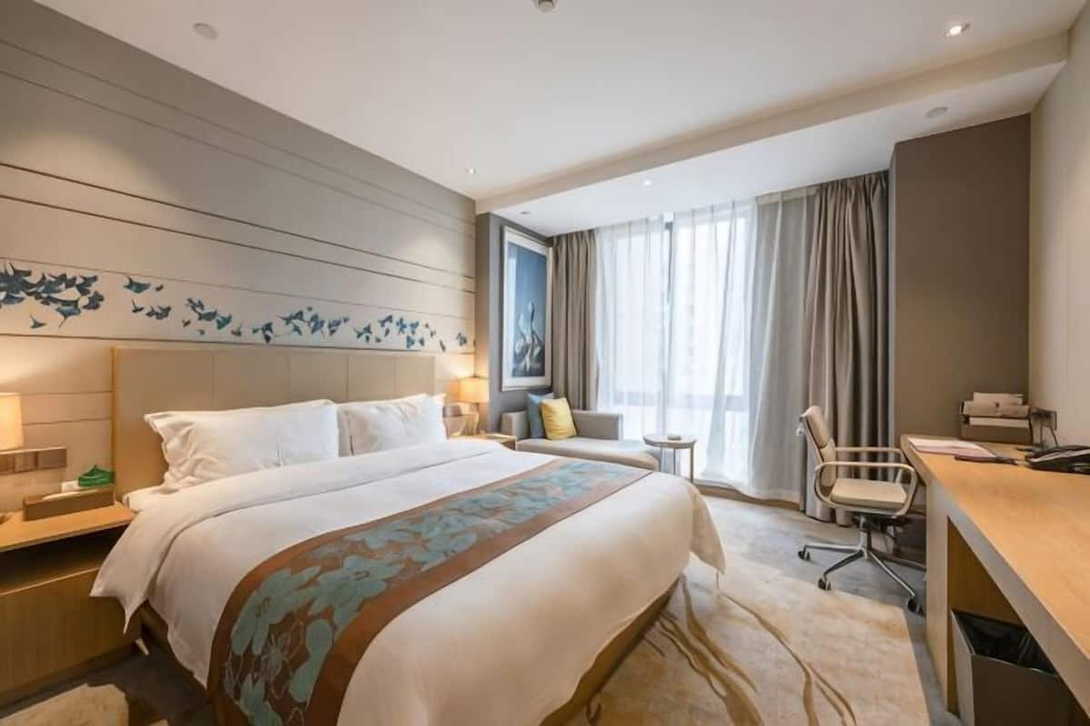 Quarto de hotel com uma cama grande, uma escrivaninha, uma cadeira, uma mesinha e uma janela com cortinas.