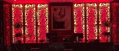 Une pièce faiblement éclairée, avec un fond rouge illuminé, une œuvre d’art encadrée accrochée au mur et un coin bar avec des tabourets.