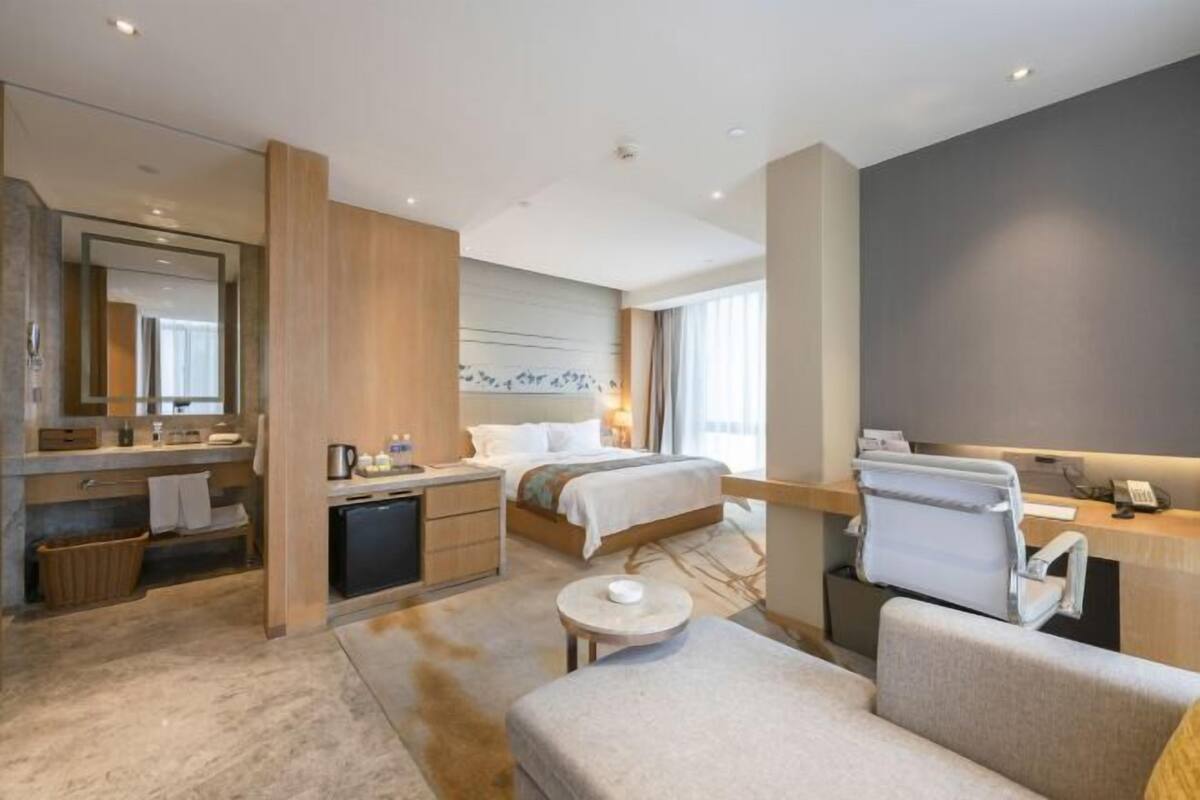 Quarto de hotel moderno com cama, escrivaninha, cadeira, mesinha e área de banheiro.