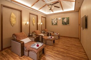 Sala de spa com piso de madeira, ventilador de teto, cadeiras de massagem fixadas na parede e obras de arte.