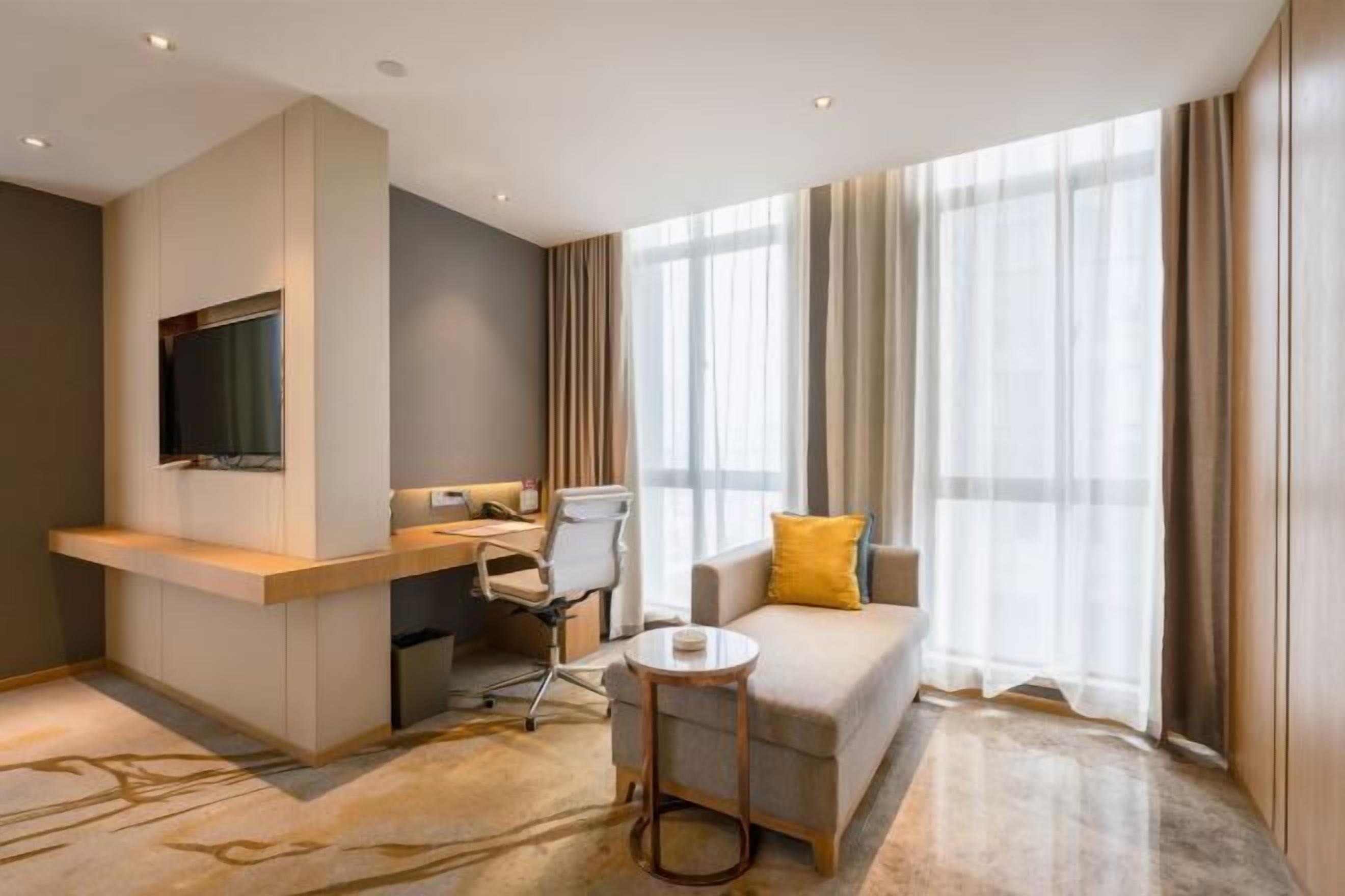 A modern hotel room with a sofa, a small round table, a desk with a chair, a TV mounted on the wall, and large windows with curtains.