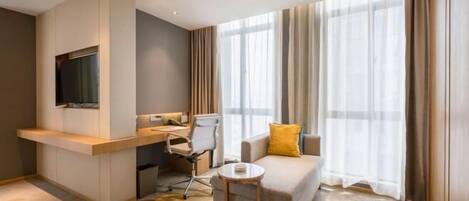 A modern hotel room with a sofa, a small round table, a desk with a chair, a TV mounted on the wall, and large windows with curtains.