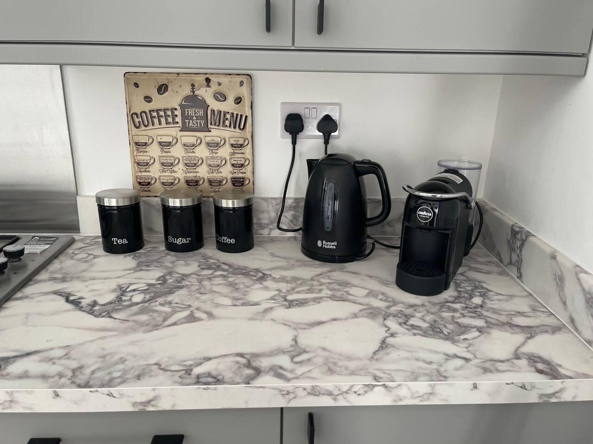 A kitchen counter with a coffee maker, a kettle, and three containers labeled tea, sugar, and coffee.