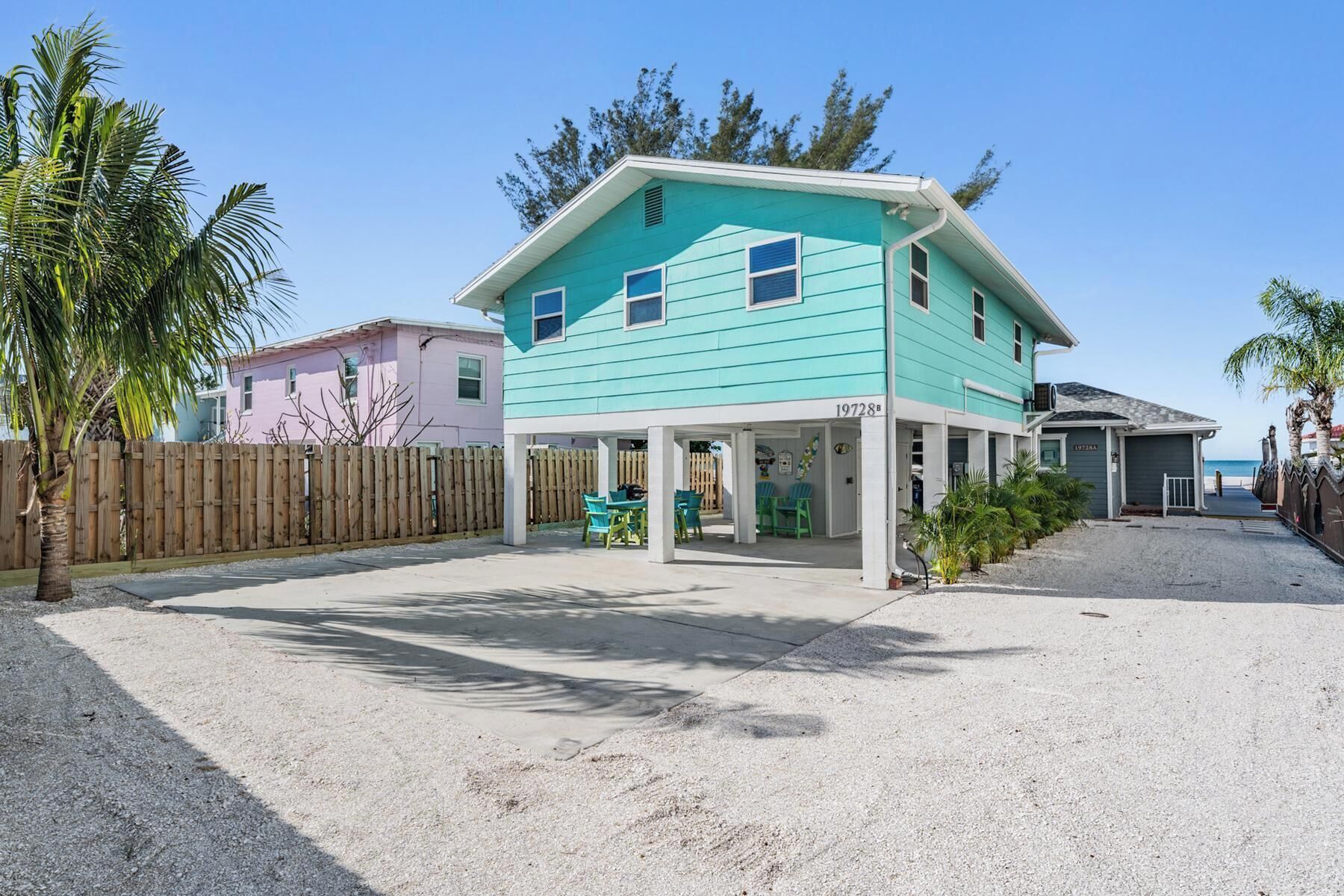 Une maison turquoise à deux étages, dotée d’un patio couvert, d’une allée en sable et d’une clôture en bois.