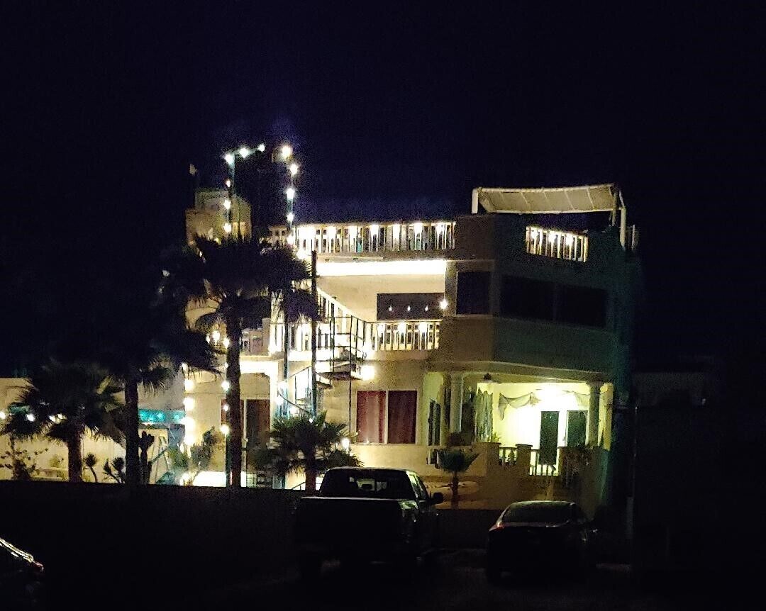 A multi-story building with balconies and outdoor lighting, parked cars in front, and palm trees.