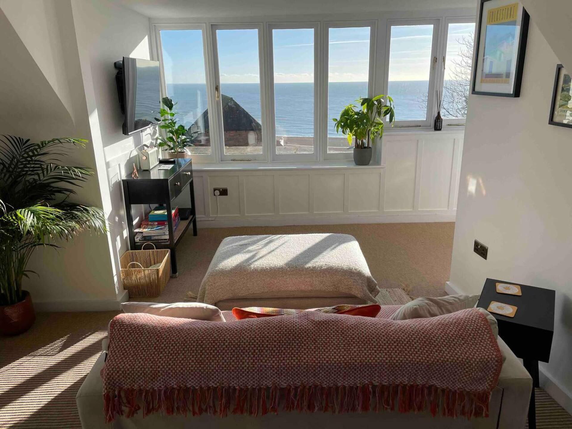 A bedroom with a large window overlooking the sea, a bed with a patterned blanket, a small desk, and a potted plant.