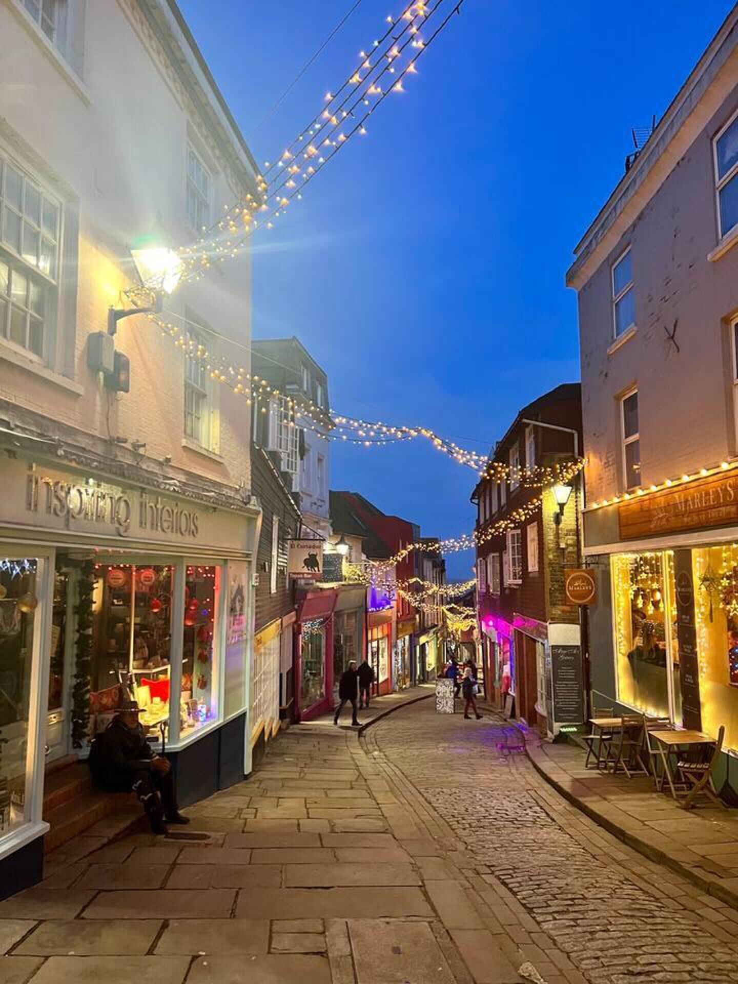 Une rue pavée bordée de boutiques et d’immeubles, illuminée de guirlandes lumineuses.