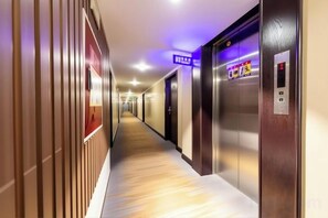 Corredor de hotel com painéis de madeira, um elevador e uma placa indicando o caminho para a recepção.