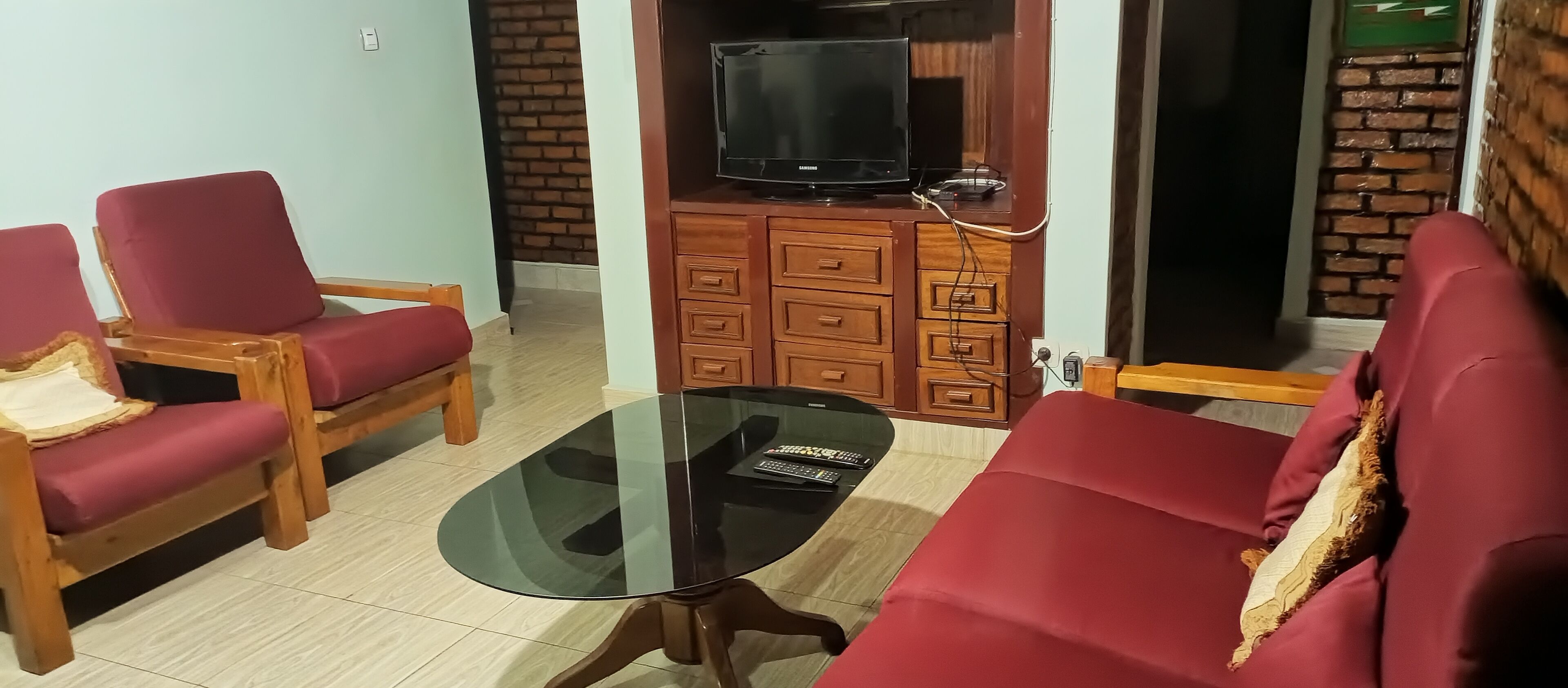 A living room with a glass-top coffee table, red sofas, wooden chairs, a TV on a wooden cabinet, and a brick wall.