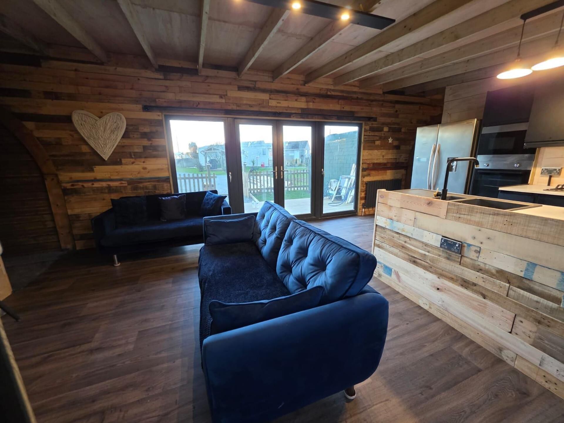 A cozy living area with a dark blue sofa, a wooden wall with a heart decoration, and a view through large glass doors.