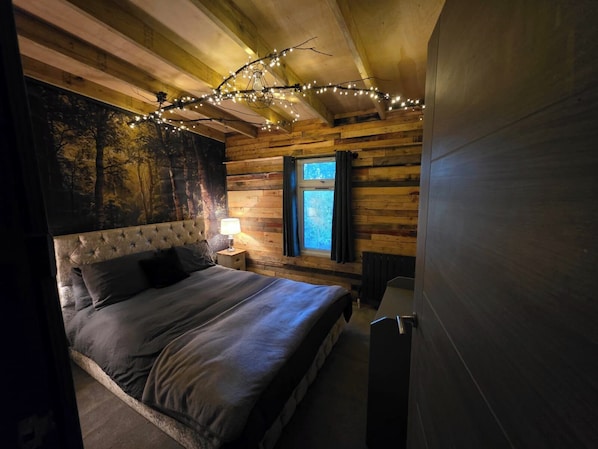 Ein Schlafzimmer mit einem großen Bett, einem Nachttisch mit Lampe, Holzwänden, einem Fenster mit Vorhängen und Lichterketten an der Decke.