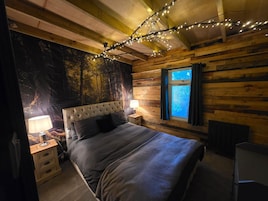 Un dormitorio con cama, mesitas de noche, lámpara, ventana con cortinas y un papel tapiz con motivos de bosque.