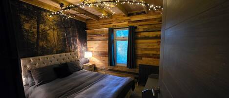 A bedroom with a large bed, a nightstand, a lamp, a window with curtains, and a wall with a forest mural.