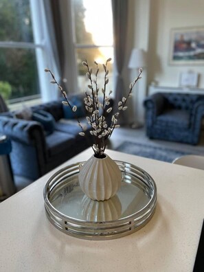 A white vase with branches on a table in a room with a sofa and a window.