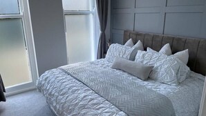 Ein Schlafzimmer mit einem großen Bett, einem grauen Kopfteil und Blick durch ein Fenster.