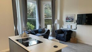 Ein modernes Wohnzimmer mit einer zentralen Kücheninsel, einem Sofa, einem Sessel und einem an der Wand montierten Fernseher.