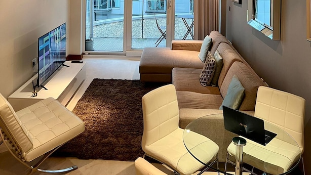 Una sala moderna con un sofá, sillones, una mesa de centro de vidrio y una computadora portátil. Hay cuadros enmarcados en la pared, un televisor de pantalla plana y un ventanal con vistas a edificios.