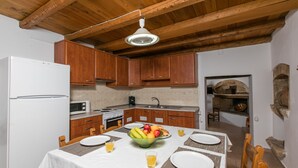 A modern kitchen with wooden cabinets, a white refrigerator, and a dining area set for four with a fruit bowl and glasses. - Pool Villa Afroditi for 10 Persons in Margarites (Mylopotamos)