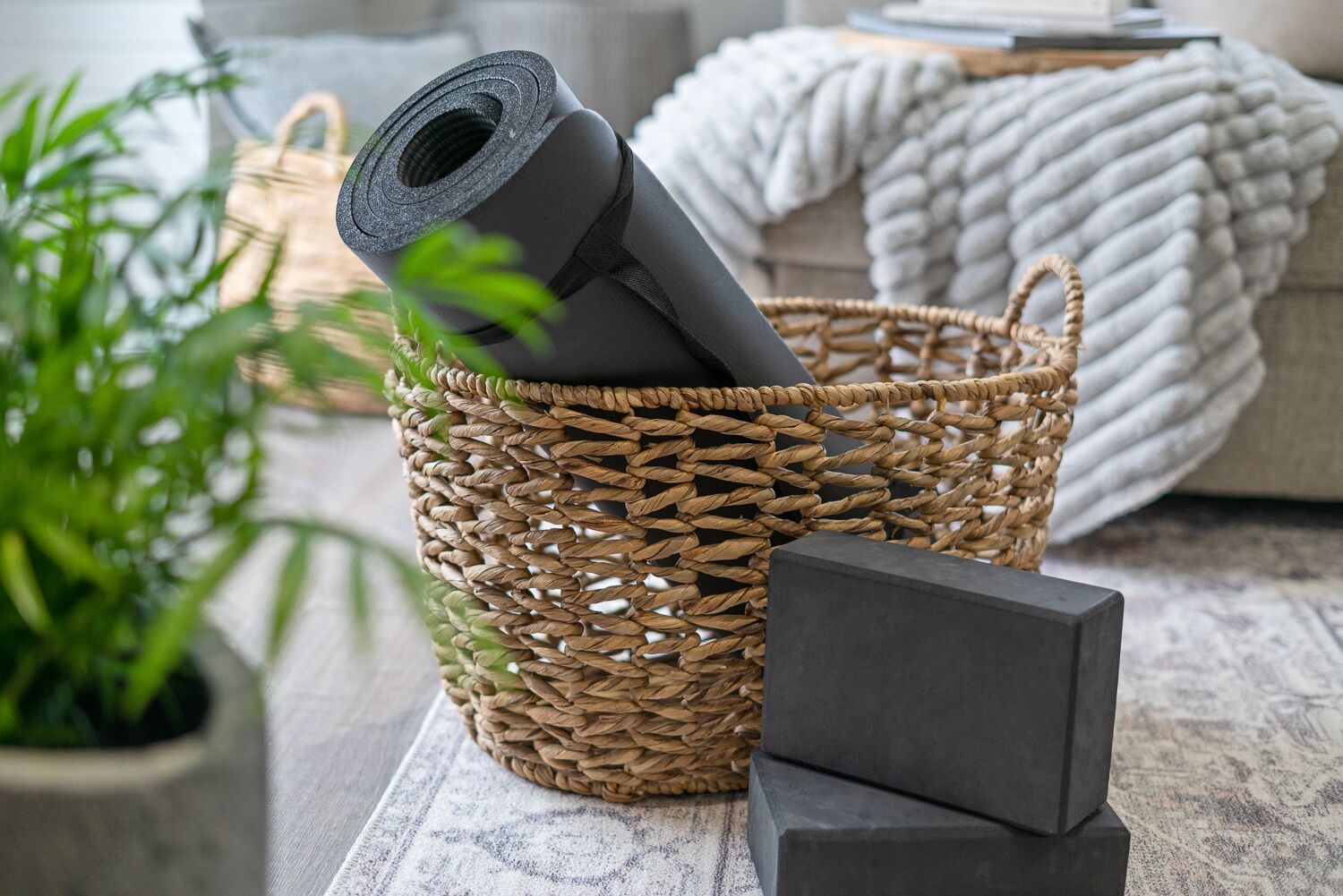 A woven basket containing a rolled yoga mat and a black block, placed on a textured surface with a plant and a textured blanket in the background.