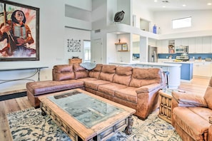 A spacious living room with a large brown leather sectional sofa, a glass-top coffee table, and a patterned rug. The room features a high ceiling and a view of the kitchen area with modern appliances.