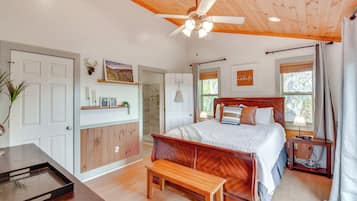 Quarto com teto de madeira, cama com cabeceira, mesa de cabeceira, um banco, cortinas na janela e uma porta.