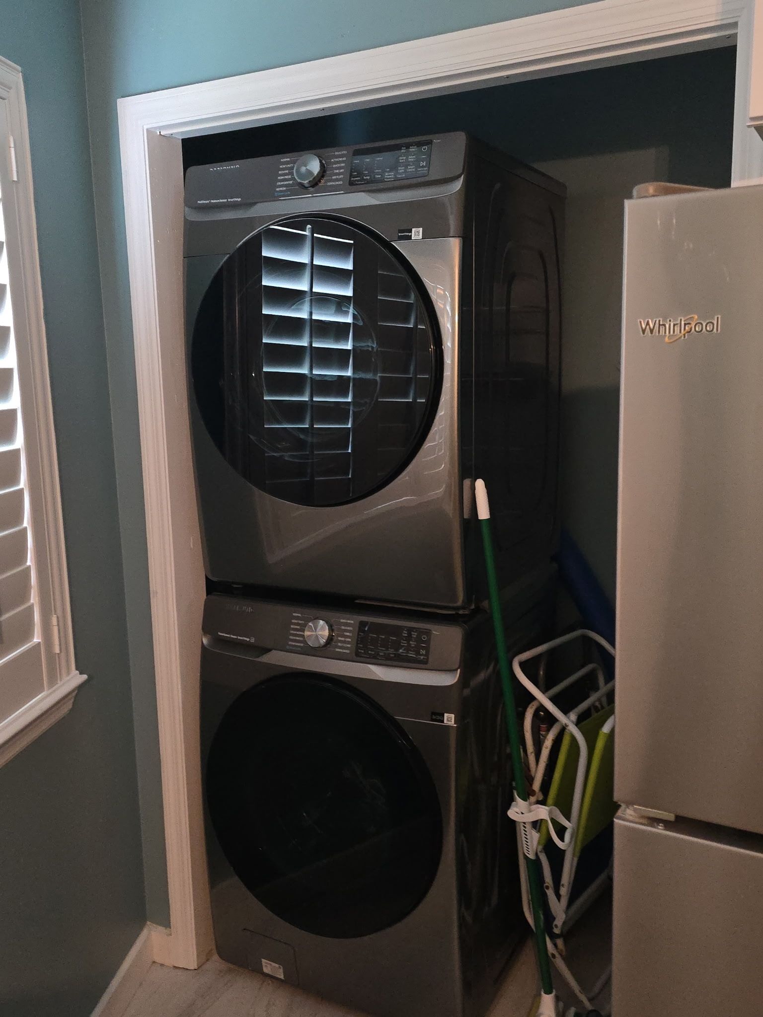 Un lavadero pequeño con lavadora y secadora apiladas, un refrigerador Whirlpool y una ventana con persianas.