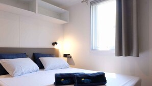 A neatly made bed with white and blue bedding, a nightstand with a lamp, and a window with curtains.
