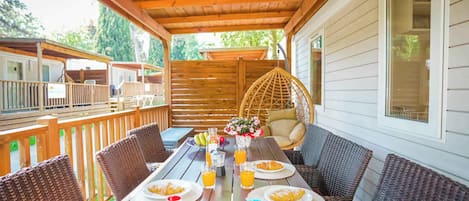 A covered outdoor dining area with wicker furniture, a wooden table set with plates, glasses, and a fruit basket.