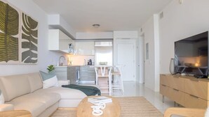Un salon moderne avec un canapé, une table basse et un téléviseur. Au fond, on aperçoit une cuisine avec des meubles blancs et un coin repas.