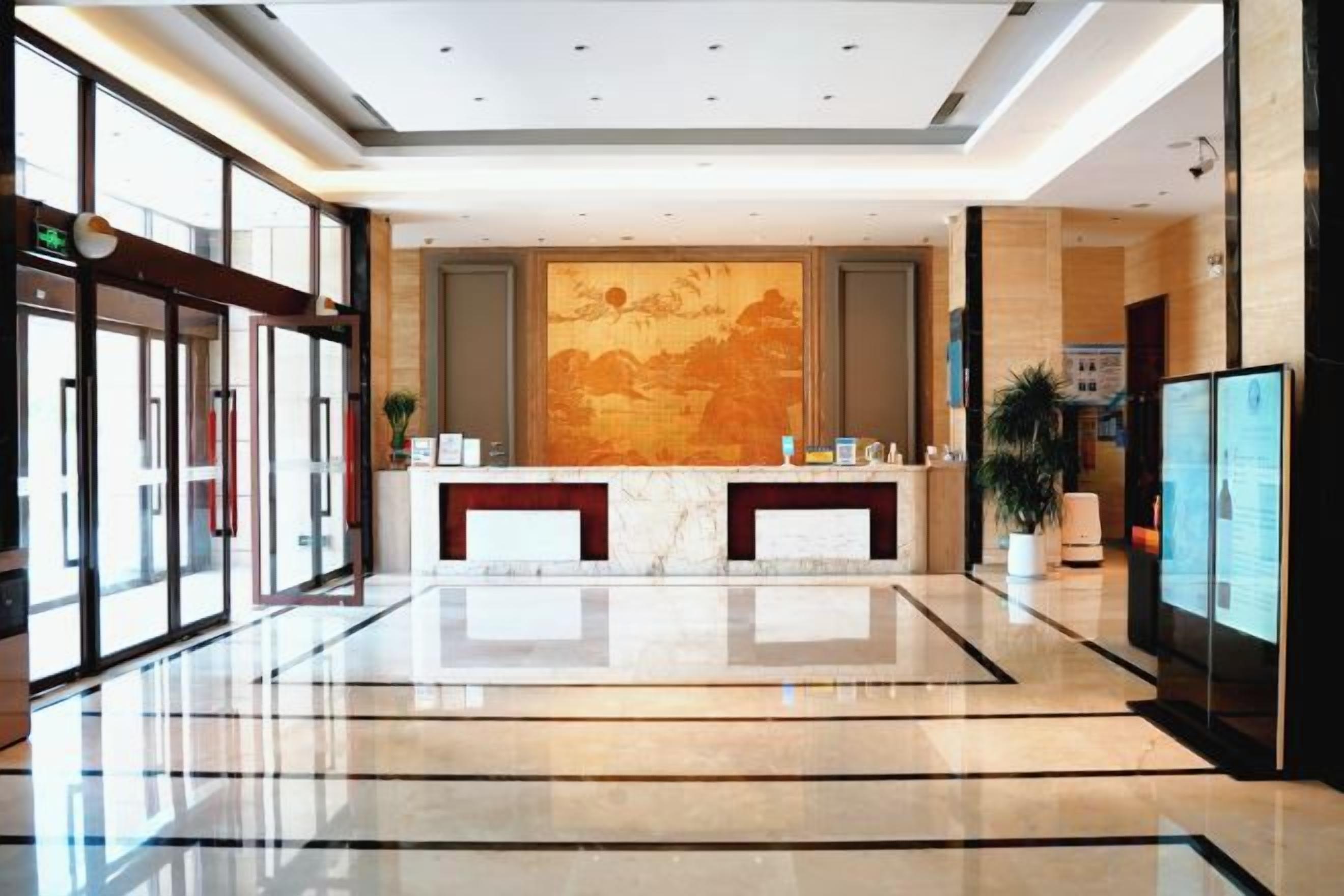 A hotel lobby with a reception desk, glass doors, a large wall mural, and a polished floor.