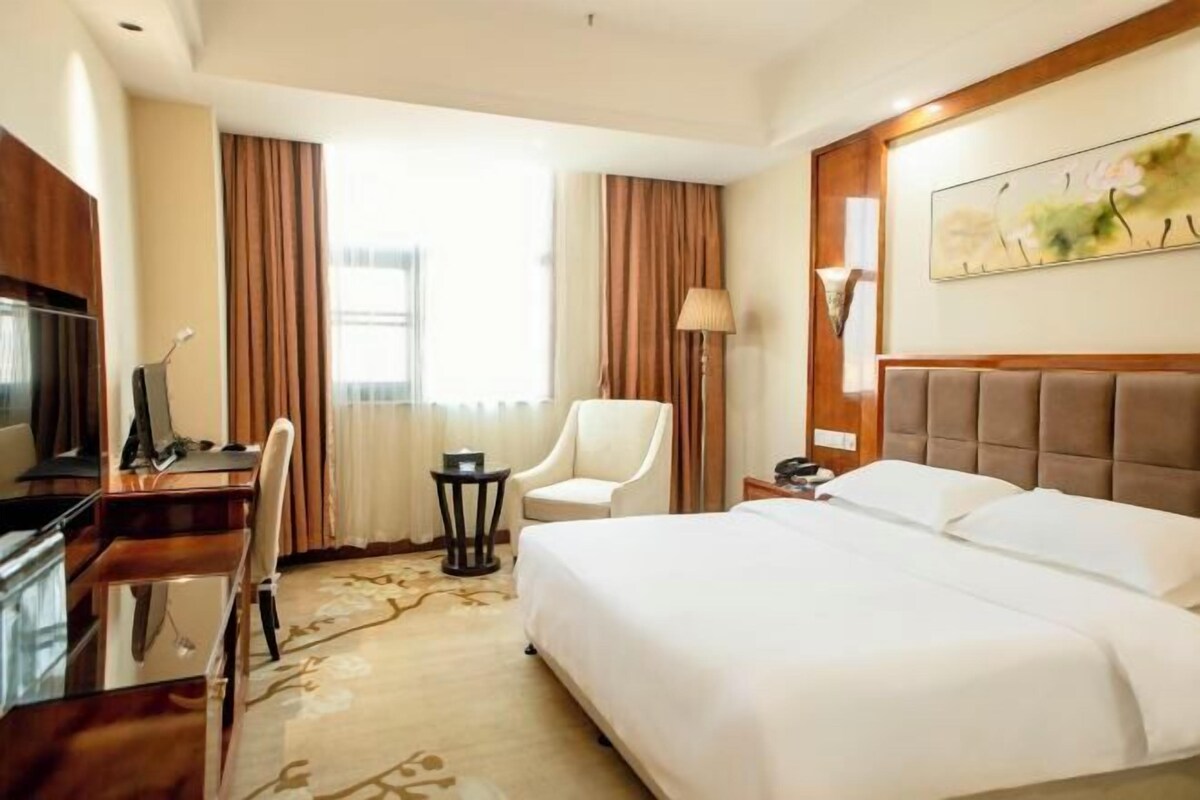 A hotel room with a large bed, a desk with a chair, a TV, a lamp, and a painting on the wall.