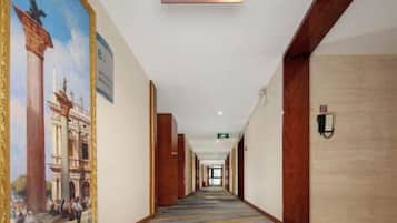 A hotel corridor with a sign indicating an elevator direction, a mural on the left wall, and a patterned carpet.