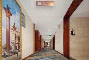 A hotel corridor with a sign indicating an elevator direction, a mural on the left wall, and a patterned carpet.