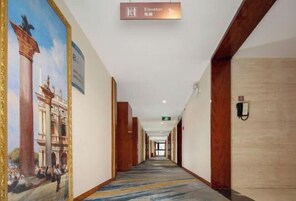 A hotel corridor with a sign indicating an elevator direction, a mural on the left wall, and a patterned carpet.
