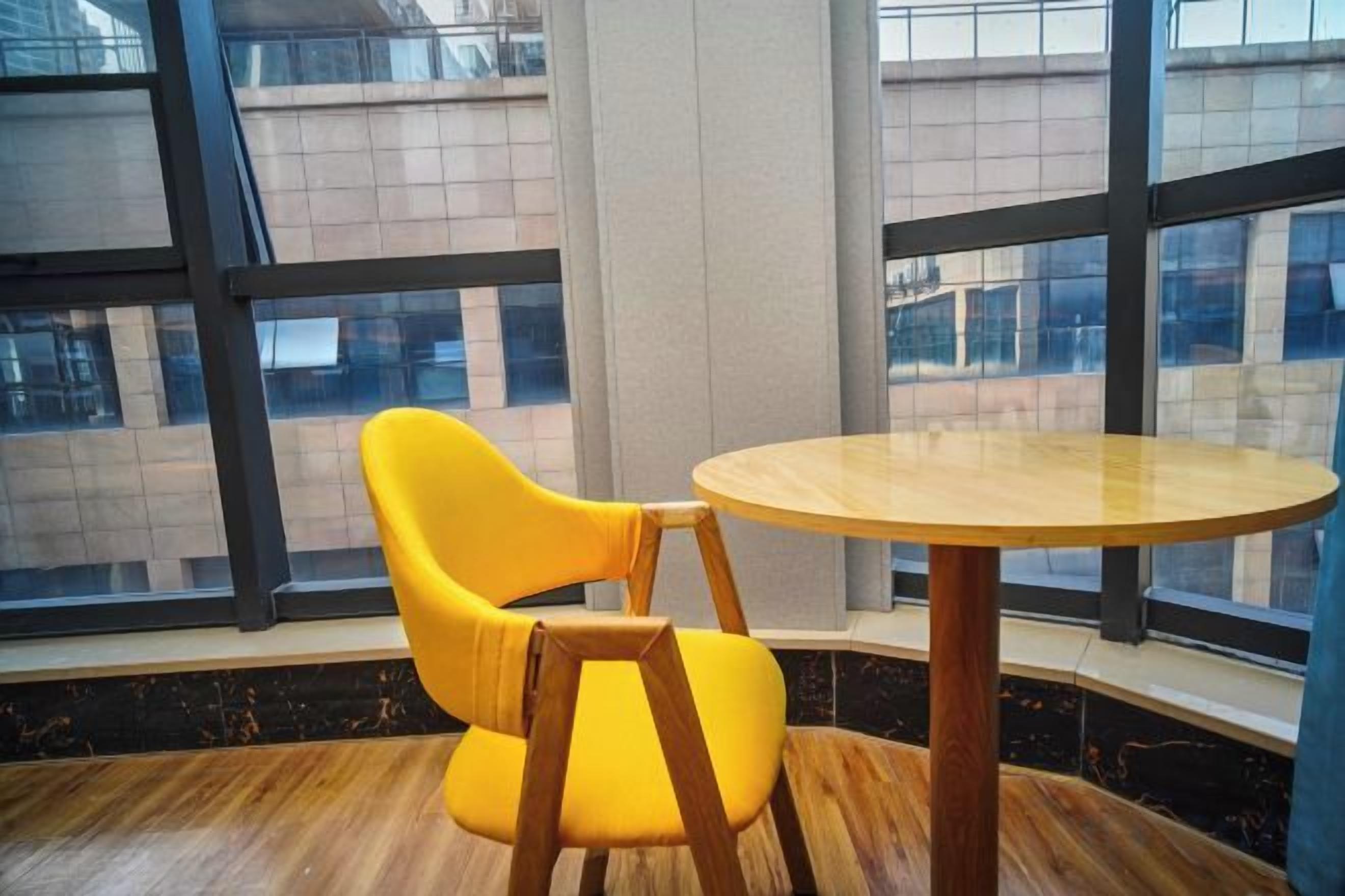 A room with a round wooden table and a yellow chair, large windows, and a view of buildings outside.