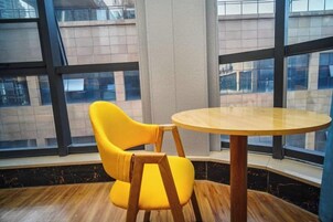 Quarto com uma mesa de madeira redonda e uma cadeira amarela, janelas amplas e vista para prédios.