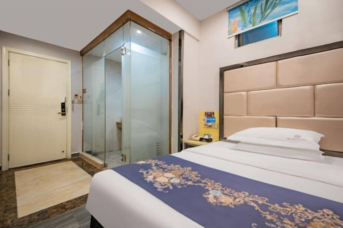 A hotel room with a bed, a glass-enclosed shower, a bedside table, and a painting on the wall.
