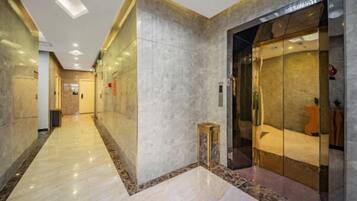 Un pasillo de hotel con piso de mármol, un ascensor de vidrio y un taburete de madera.