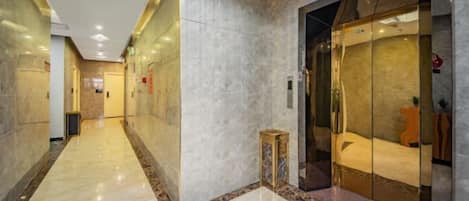 A hotel corridor with a marble floor, a glass elevator, and a small wooden stool.