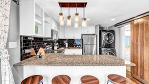 Una cocina con encimeras de granito, armarios blancos, un refrigerador de acero inoxidable, horno empotrado y una barra de madera con taburetes.