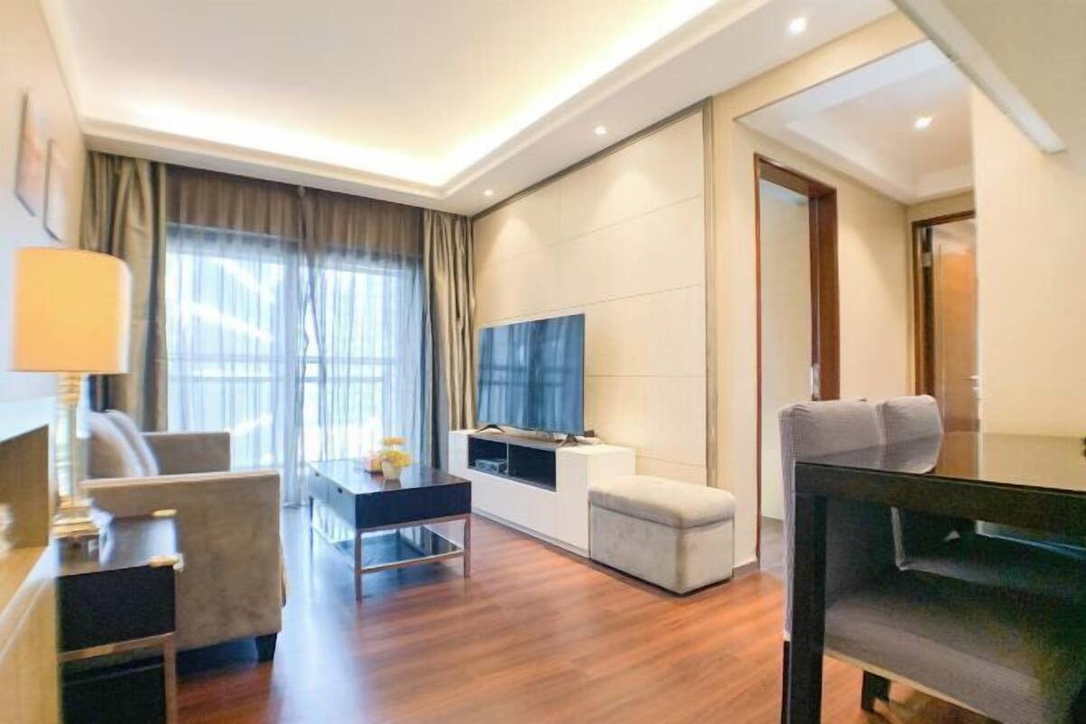 A modern hotel room with a wooden floor, a flat-screen TV, a sofa, a desk, and a small ottoman.
