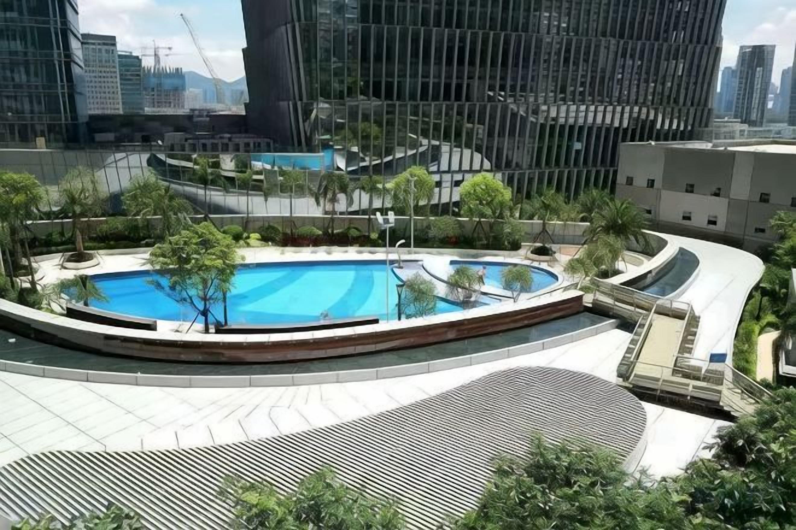 A rooftop swimming pool with a circular design, surrounded by greenery and modern buildings in the background.