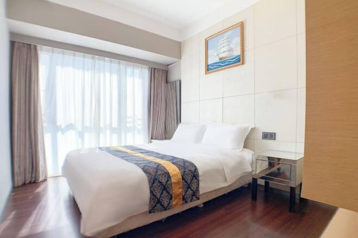 A hotel room with a bed, a bedside table, a painting on the wall, and a window with curtains.