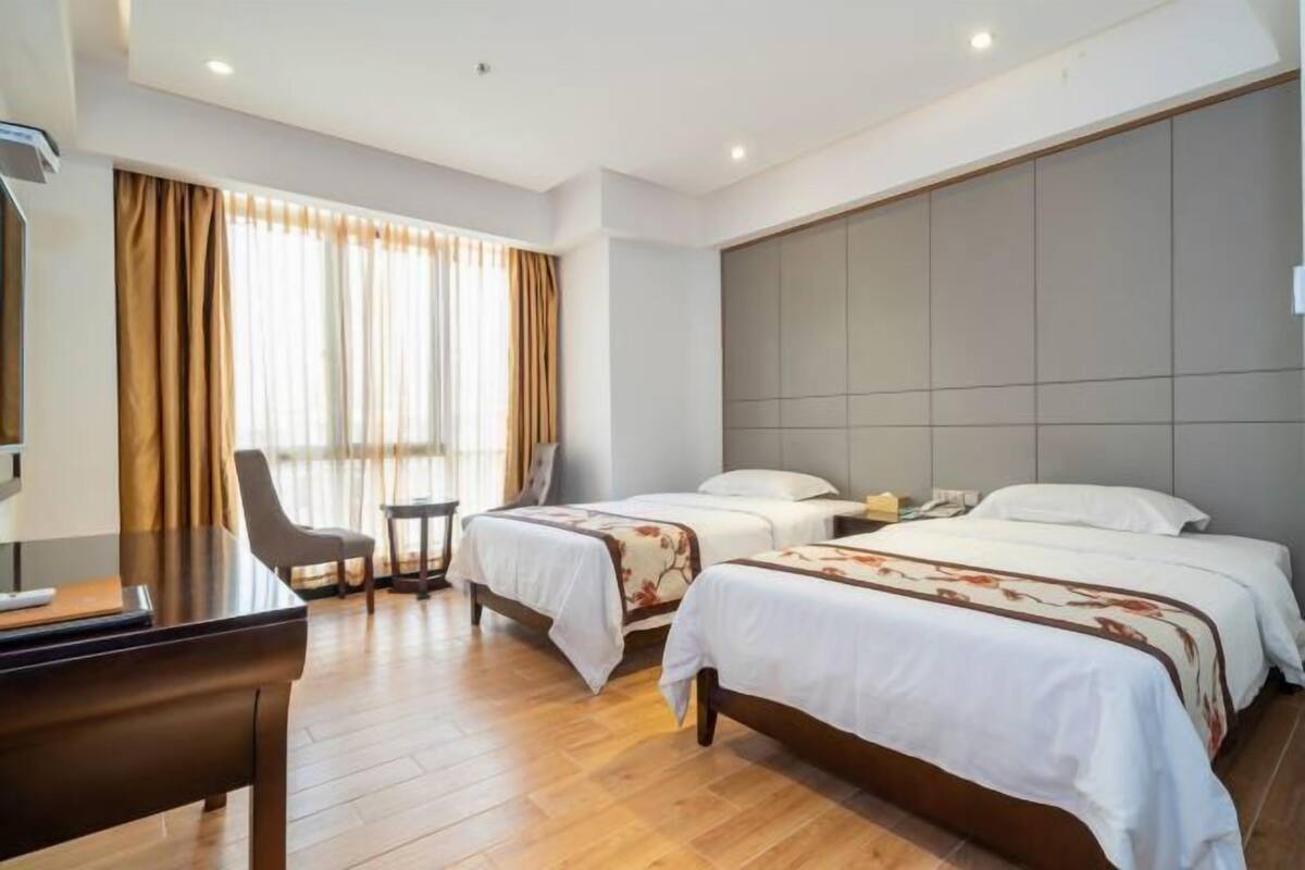 Quarto de hotel com duas camas, uma escrivaninha, uma cadeira e uma janela grande com cortinas.