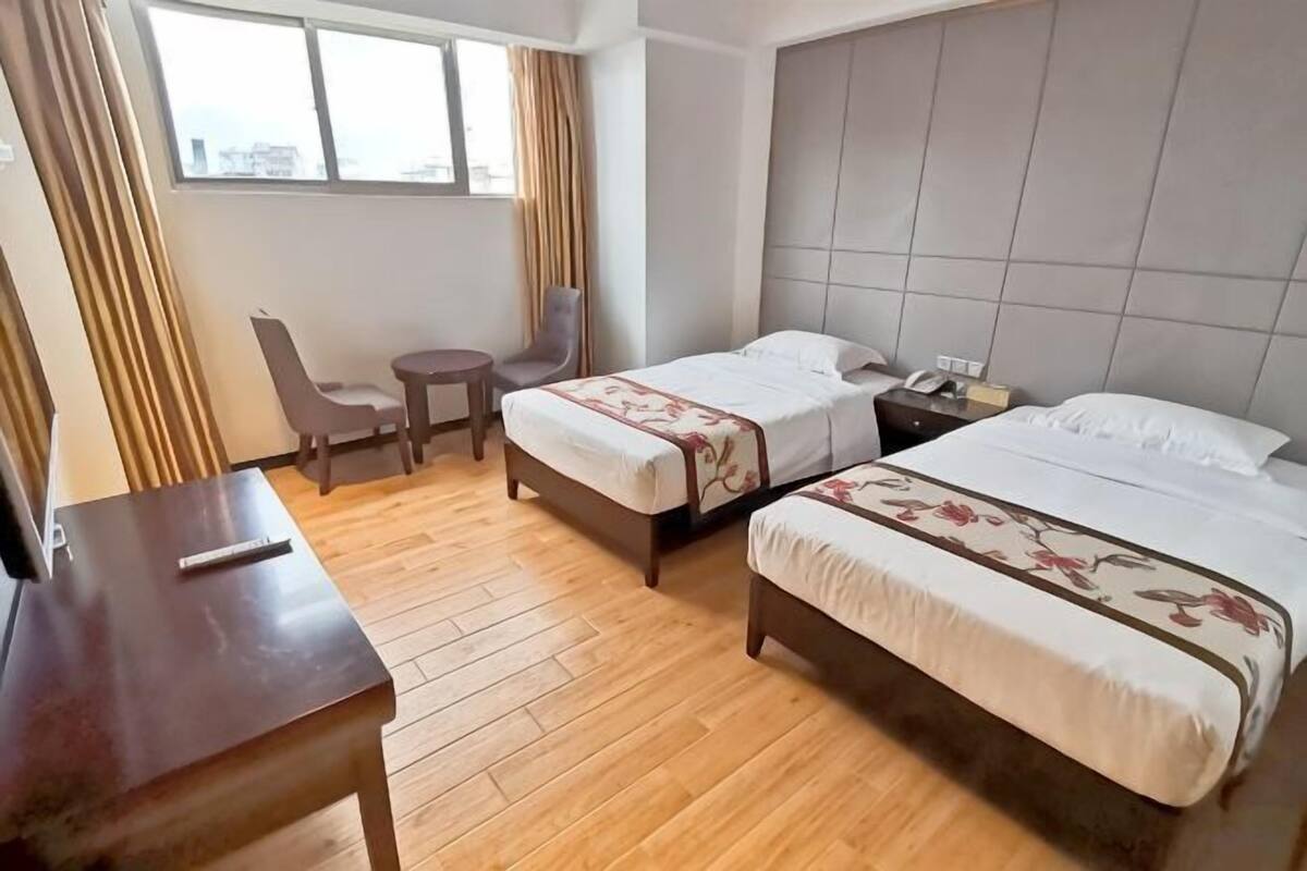 Quarto de hotel com duas camas, uma escrivaninha, uma cadeira, uma mesinha e uma janela com cortinas.
