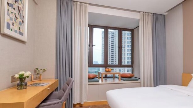 Ein Hotelzimmer mit Schreibtisch, Stuhl, Bett, Fenster mit Vorhängen und Blick auf Gebäude.