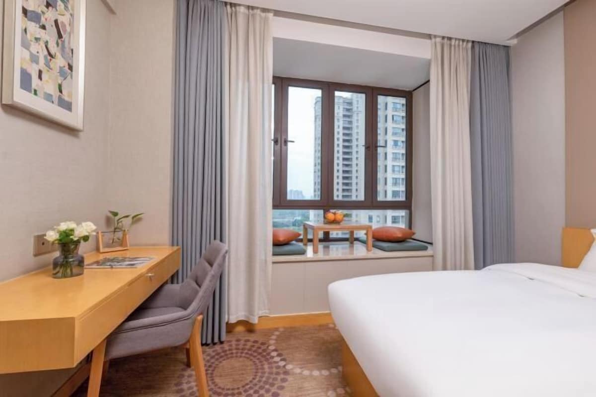 Ein Hotelzimmer mit Schreibtisch, Stuhl, Bett, Fenster mit Vorhängen und Blick auf Gebäude.