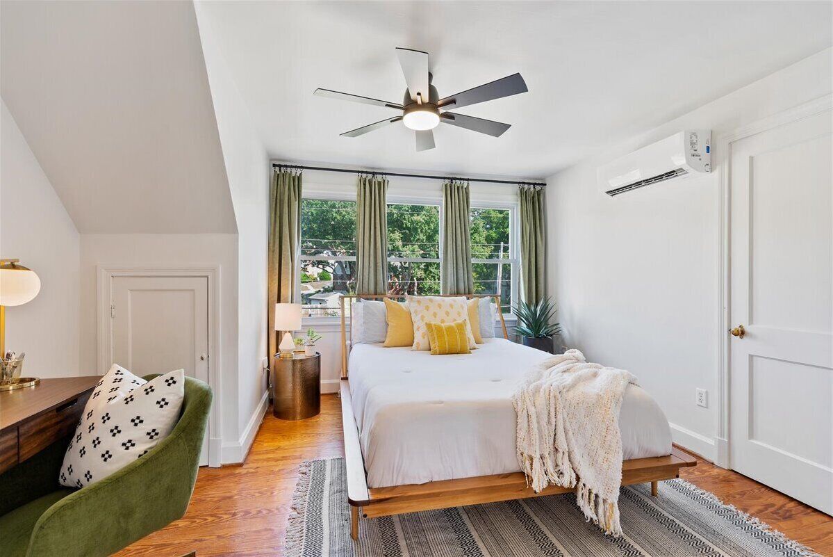 A bedroom with a bed, a ceiling fan, a window with curtains, a bedside table, and a green armchair.