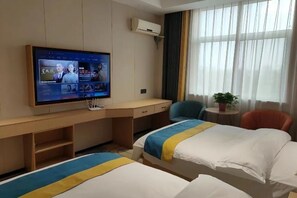 Ein Hotelzimmer mit Fernseher an der Wand, einem Schreibtisch, zwei Betten mit blauen und gelben Tagesdecken, einem Sessel, einem kleinen Tisch mit Pflanze und einem Fenster mit Vorhängen.